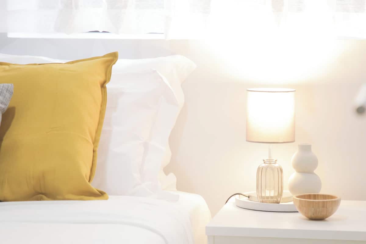 Un letto rifatto con un cuscino giallo, un comodino bianco con una lampada e un vaso.