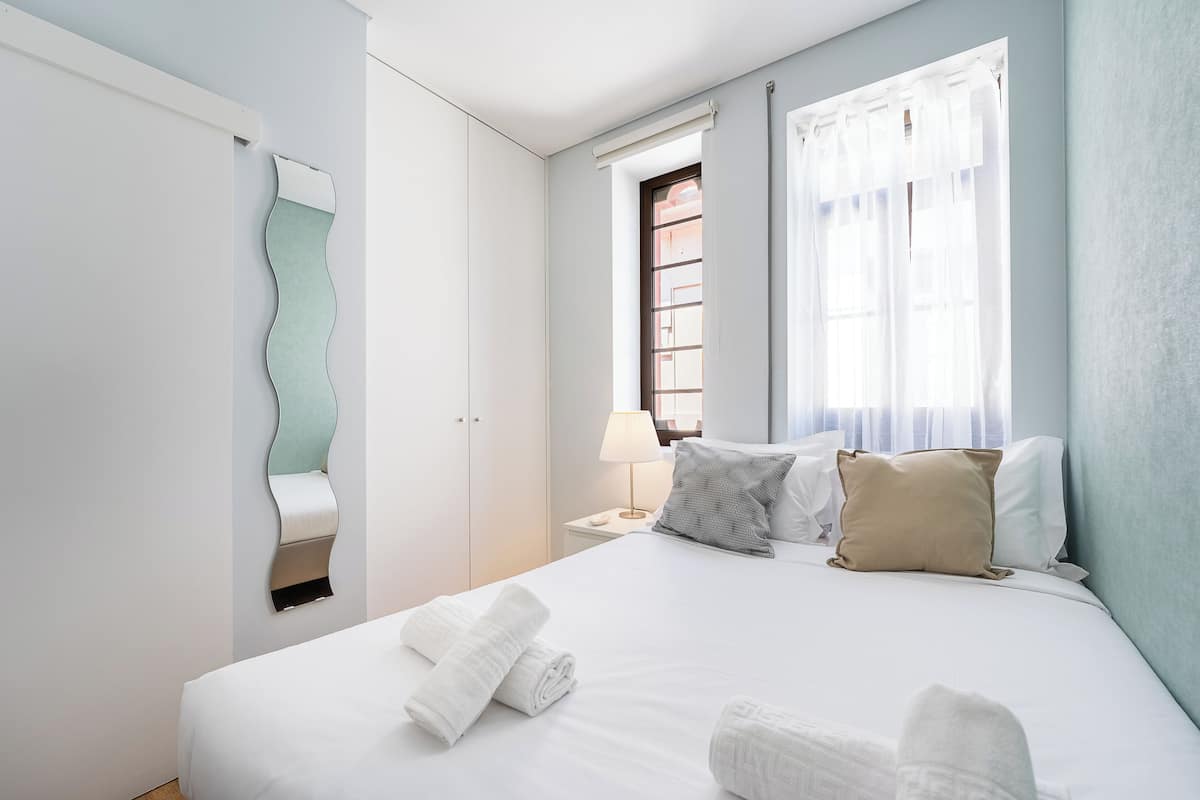 Un letto rifatto con lenzuola bianche, una lampada da comodino e una finestra con tende.