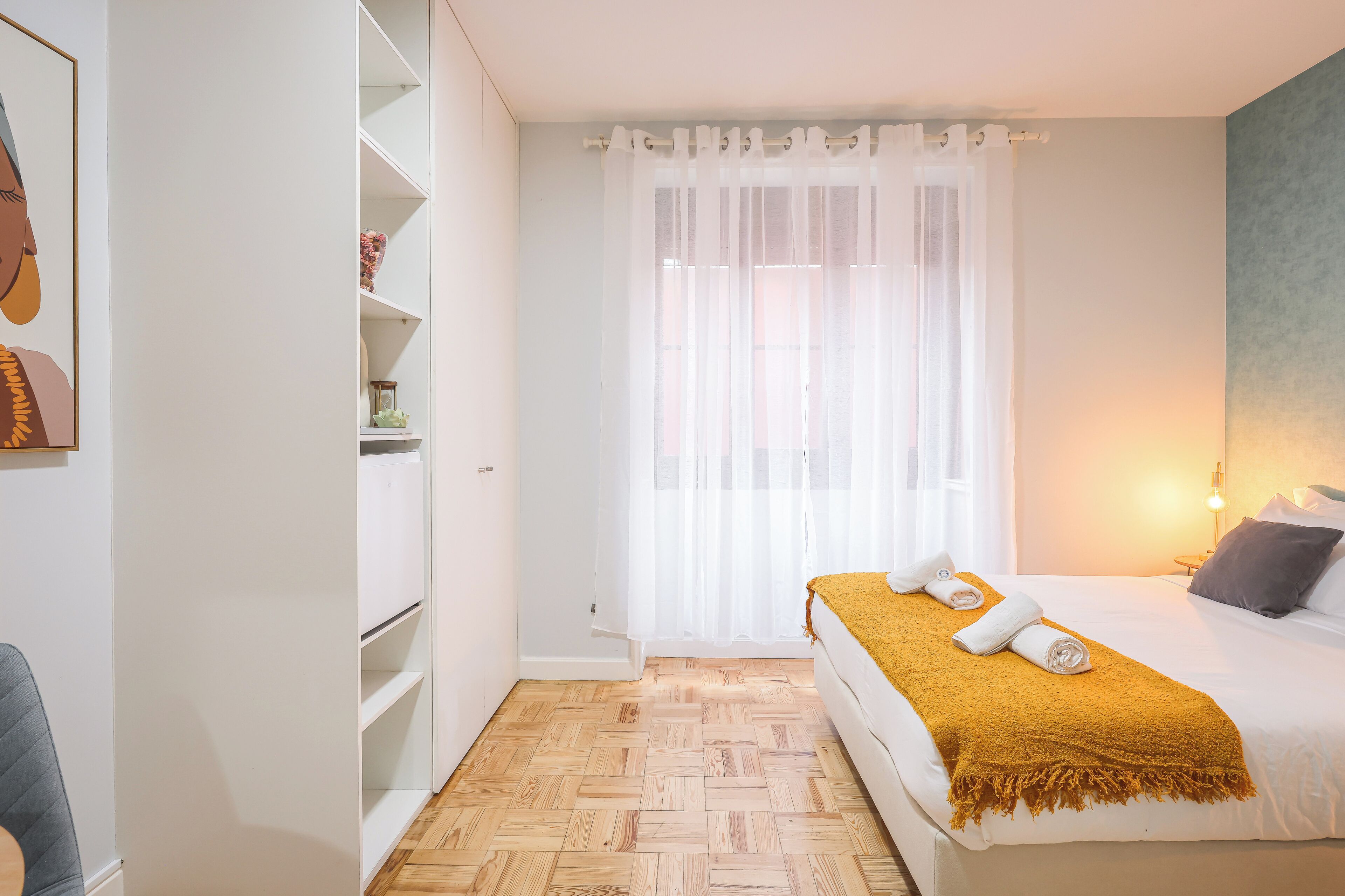 A bedroom with a bed, a window with sheer curtains, a bedside table with a lamp, and a wardrobe.