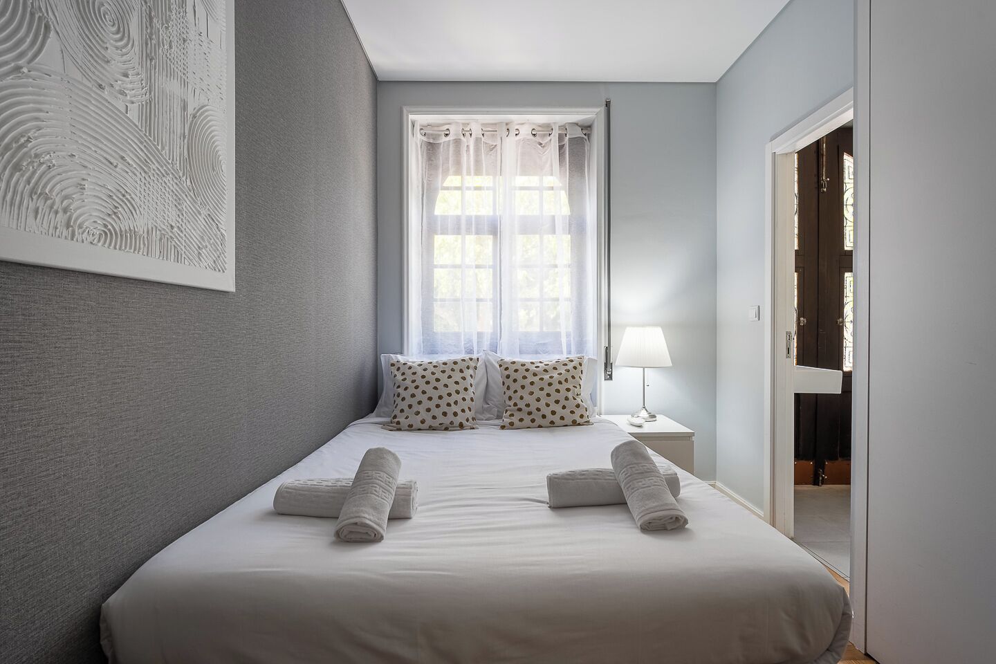 A bedroom with a bed, pillows, a nightstand, a lamp, a window with curtains, and a door.