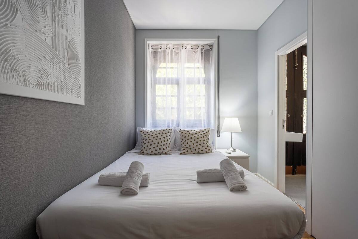 Una camera da letto con un letto, cuscini, un comodino, una lampada, una finestra con tende e una porta.