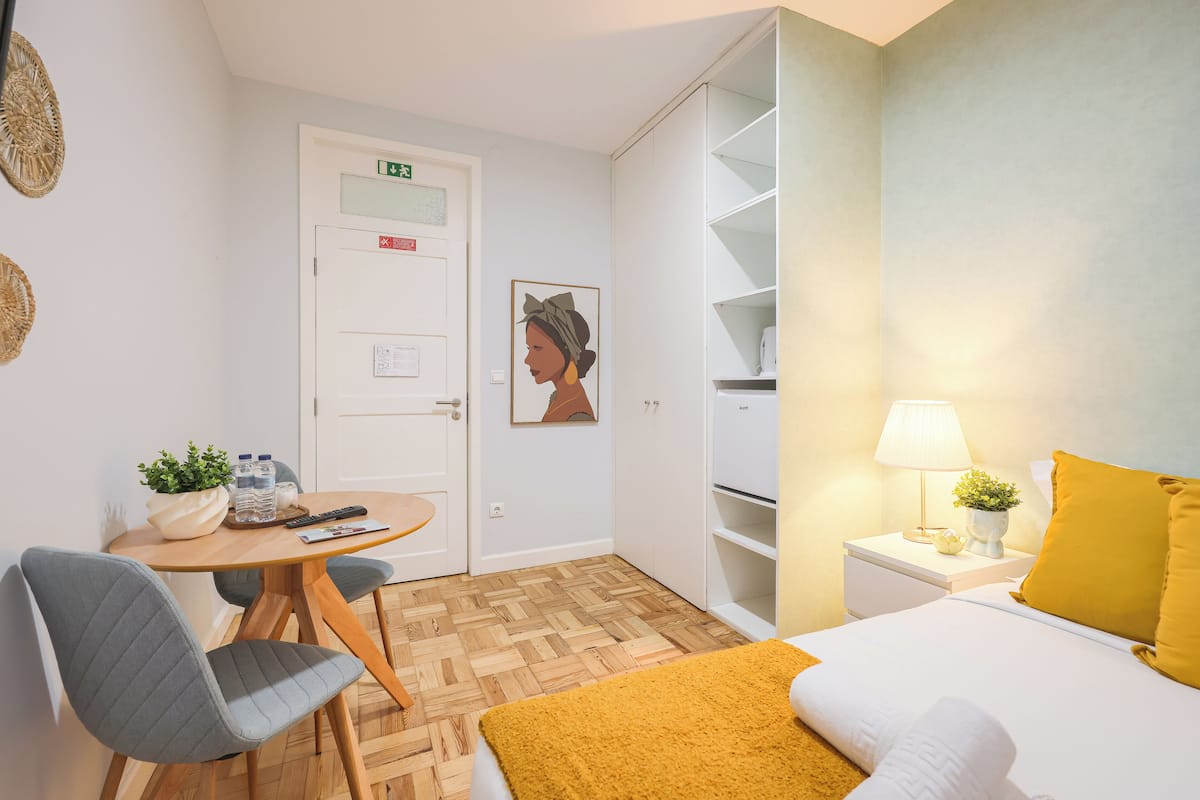Una camera d'albergo con un letto, un tavolino rotondo con sedie, una piccola pianta, un comodino con una lampada e un armadio.
