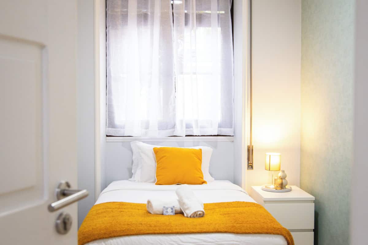 Una camera d'albergo con un letto, una finestra con tende leggere, una lampada da comodino e una porta.