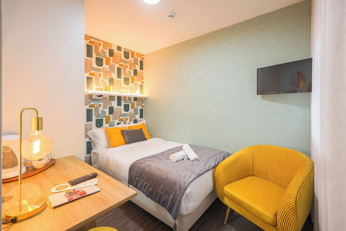 Una camera d'albergo con un letto, un comodino con una lampada, una sedia e una TV a parete.