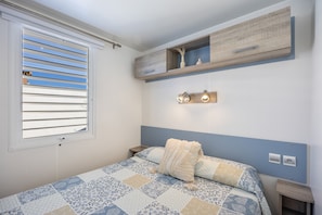 A bedroom with a bed, a window with blinds, a bedside table, a nightstand, a wall-mounted shelf, and a wall-mounted light fixture.