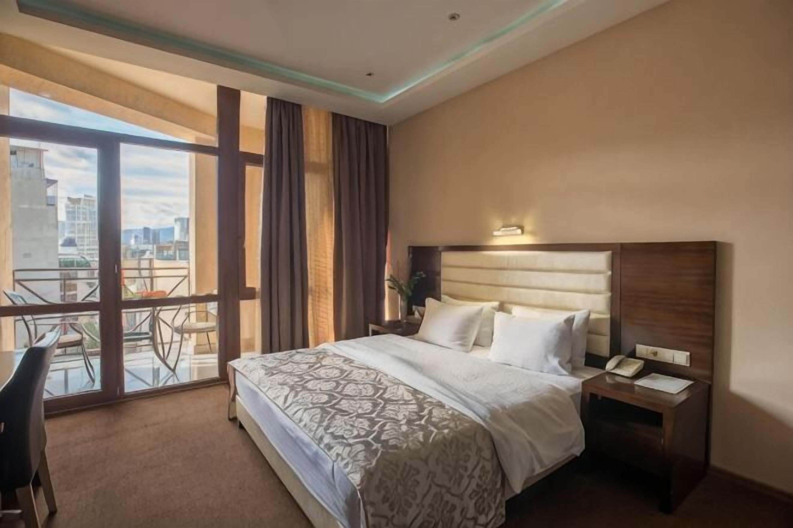 A hotel room with a large bed, a bedside table with a phone, a chair, and a balcony with a city view.