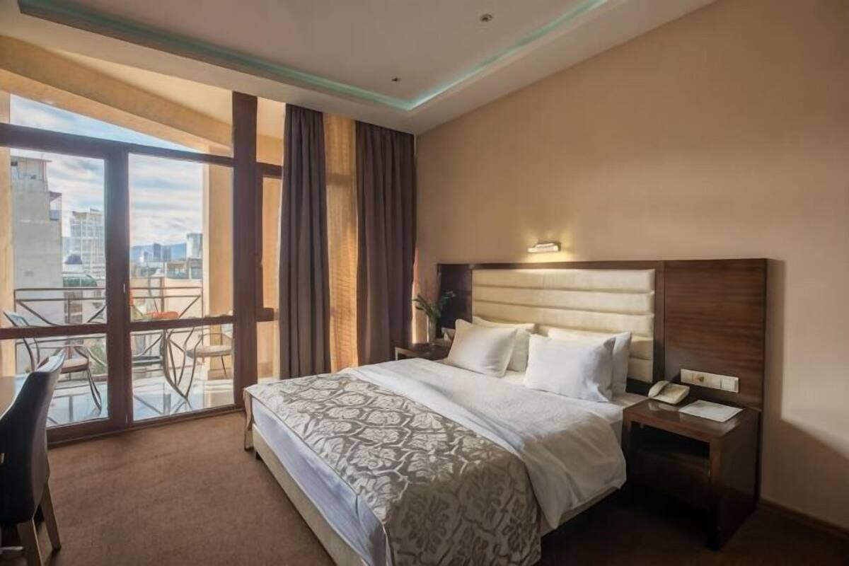 Ein Hotelzimmer mit einem großen Bett, einem Nachttisch mit Telefon, einem Sessel und einem Balkon mit Blick auf die Stadt.