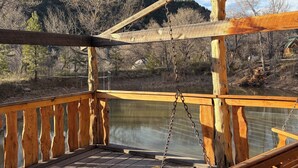 Une terrasse en bois avec un fauteuil suspendu, une balustrade et une vue sur un plan d’eau et des arbres.