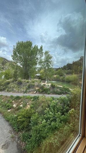 A view through a window of a natural landscape with trees, a river, and a hilly terrain.