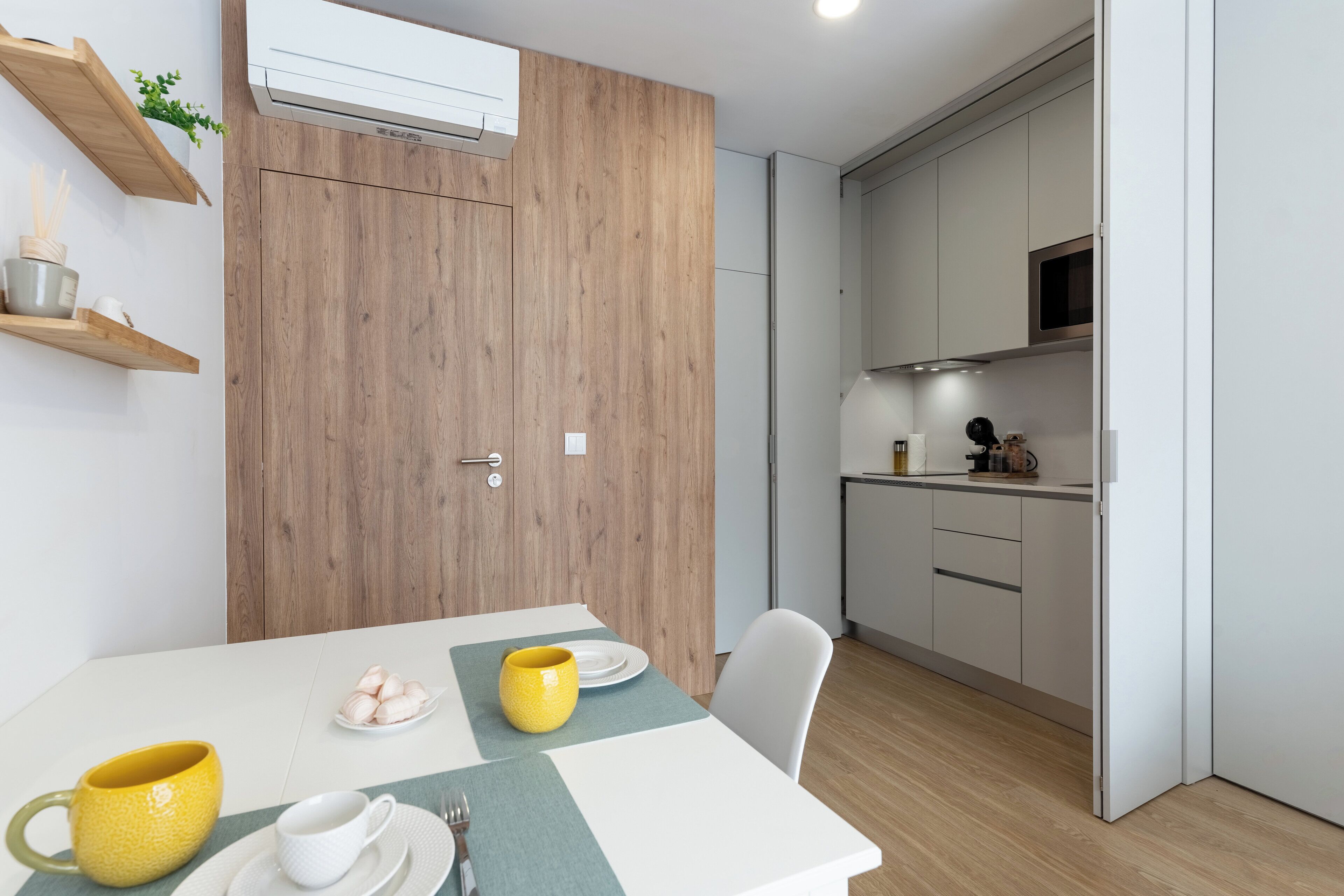 Eine moderne Küche mit weißen Schränken, einer Mikrowelle und einer kleinen Arbeitsfläche. Es gibt eine Holztür mit Griff und eine kleine Pflanze an der Wand.