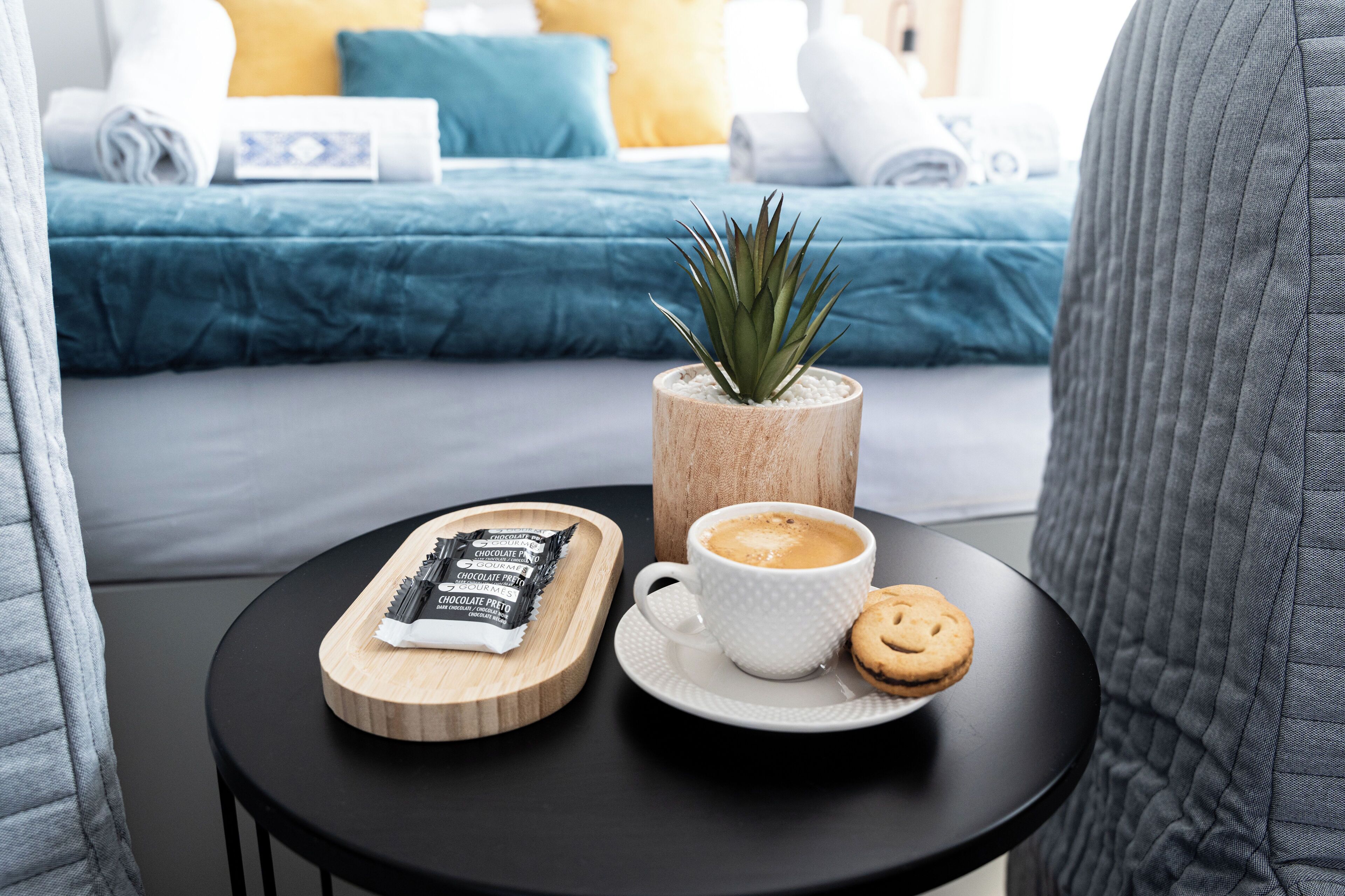 A neatly arranged breakfast table with a cup of coffee, a cookie, and chocolate bars on a tray, set against a backdrop of a bed with blue bedding and a potted plant.
