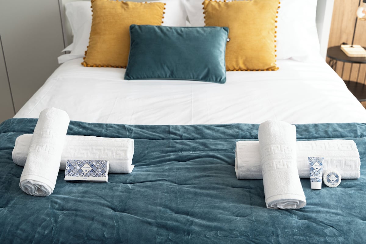 A neatly made bed with white and teal bedding, two towels, and a toiletry set on a teal bedspread.
