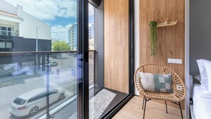 Une chambre moderne dotée d’une grande porte vitrée donnant sur un balcon, d’un fauteuil en osier et d’une petite étagère sur laquelle est posée une plante.
