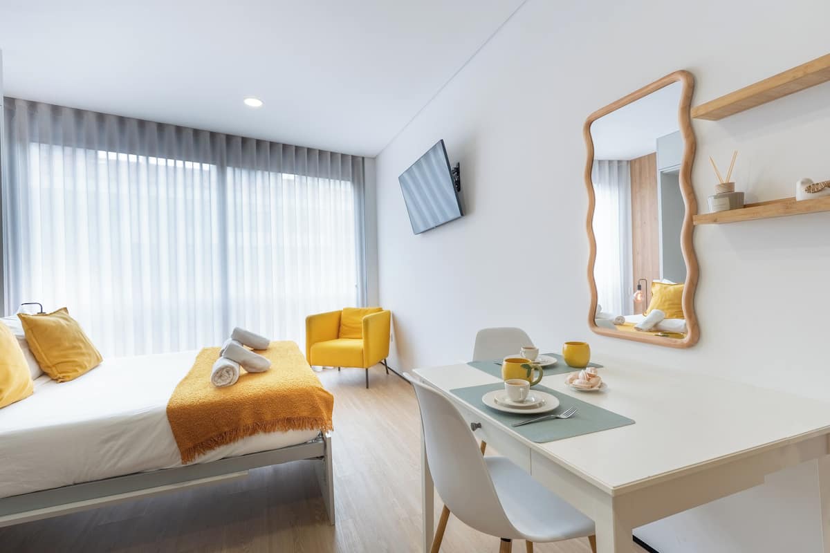 A modern hotel room with a bed, a desk, a chair, a mirror, and a window with curtains.