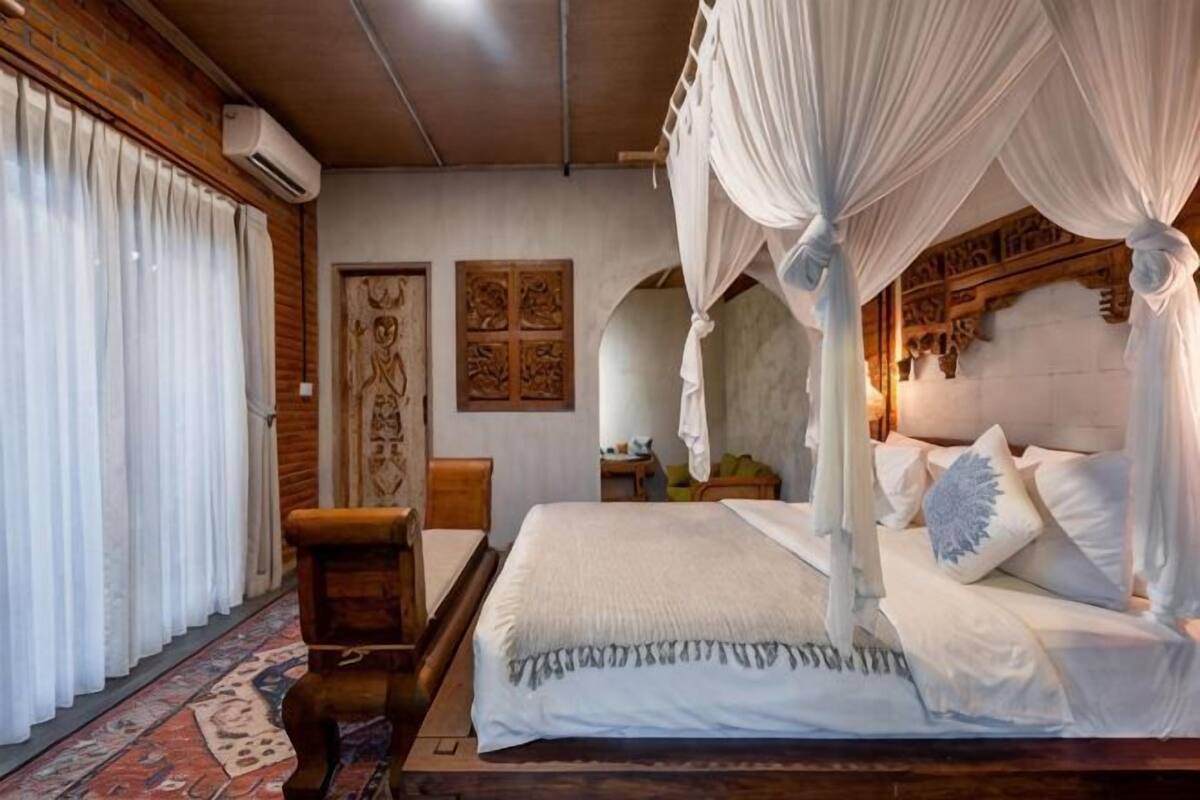 A bedroom with a four-poster bed, a wooden bench, a patterned rug, and a wall with a carved wooden panel.