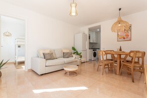 A living room with a sofa, a dining table with chairs, a hanging light fixture, and a kitchen area in the background.