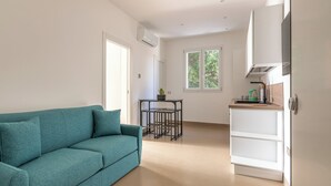 Un soggiorno moderno con un divano color petrolio, un piccolo tavolo nero con una pianta e un'area cucina bianca con pensili e una finestra.