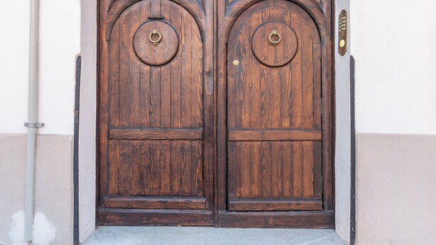 Una coppia di porte in legno con un arco in metallo sopra, incassate in un edificio con una parete esterna chiara.