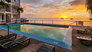 Ein Pool ohne Ende mit Liegestühlen, Blick auf den Sonnenuntergang über dem Meer und ein Gebäude mit Balkonen.