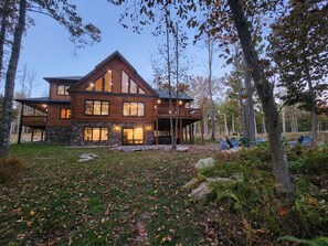 A large log cabin with multiple windows and a balcony, surrounded by trees and a grassy yard.