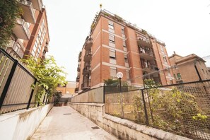 A multi-story residential building with balconies, a concrete pathway, and a metal fence.