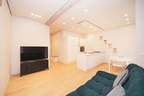 Ein modernes Wohnzimmer mit Flachbildfernseher, einem grünen Sofa und einer Küchenzeile mit weißen Schränken und einem Esstisch.