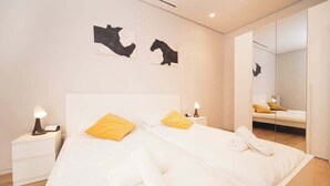 Moderni hotellihuone, jossa on kaksi sänkyä, yöpöytä lampun kanssa, suuri peili ja taideteos seinällä.