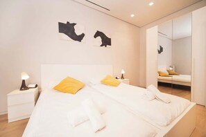 A modern hotel room with two beds, a nightstand with a lamp, a large mirror, and artwork on the wall.