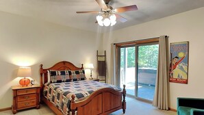 A bedroom with a wooden bed, a ceiling fan, a nightstand with a lamp, a green armchair, and a large window with curtains. - Black Pines Lodge (Swanton)