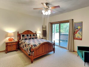 Un dormitorio con una cama de madera, ventilador de techo, mesita de noche con lámpara, sillón verde y una ventana grande con cortinas.