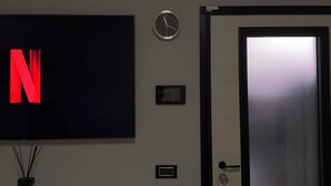 Habitación de hotel con televisor de pantalla plana mostrando el logotipo de Netflix, un reloj de pared, una puerta con panel de vidrio esmerilado y un equipo de aire acondicionado pequeño.