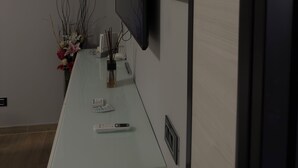 Una habitación moderna con una televisión de pantalla plana montada en la pared, una mesita con un control remoto, un florero con flores y un reloj en la pared.