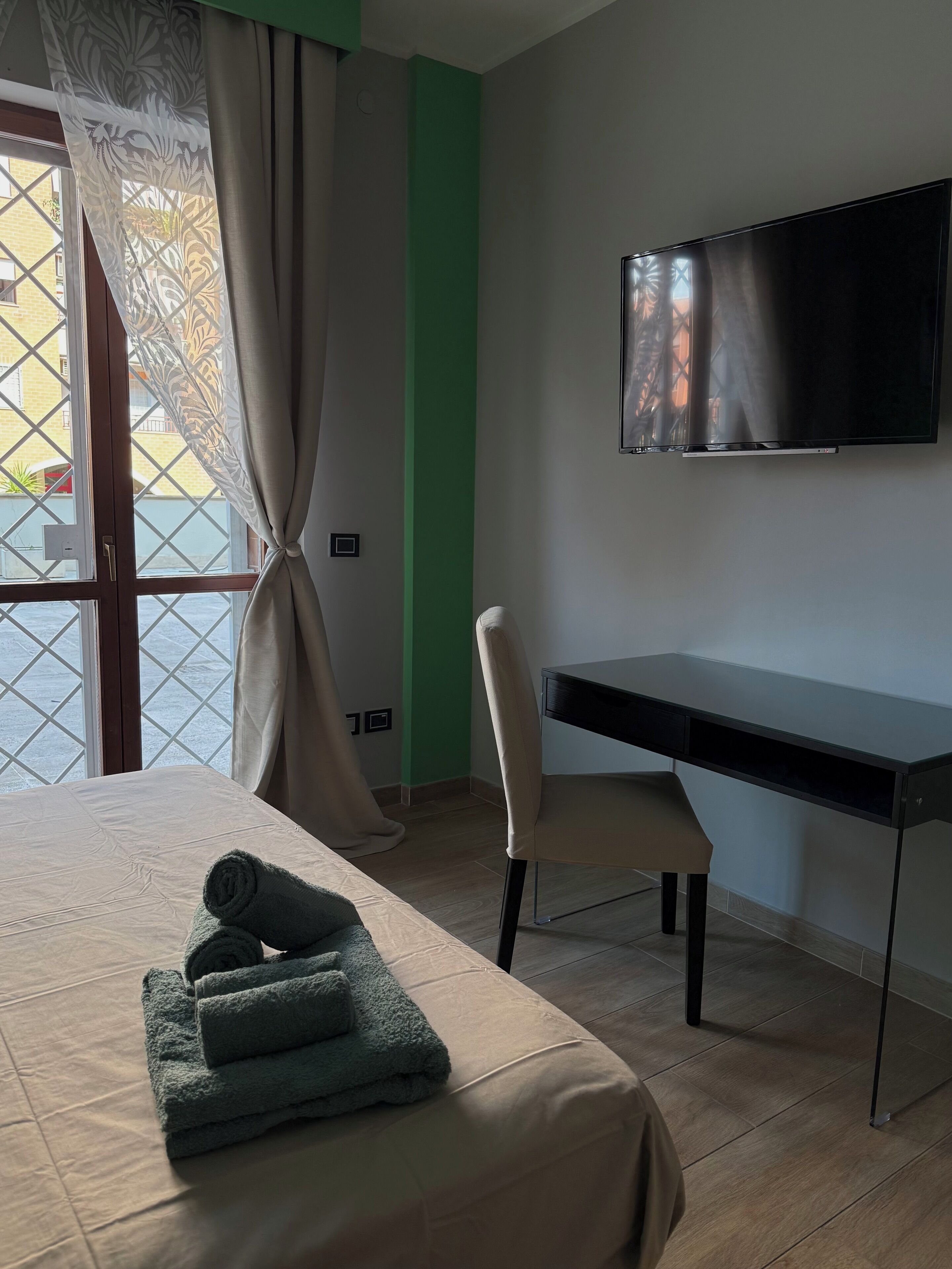Quarto de hotel com cama, escrivaninha, cadeira, televisão e janela com cortinas.