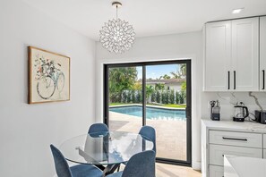 Une cuisine moderne avec des meubles blancs, un coin repas doté d’une table en verre et une vue sur une piscine.