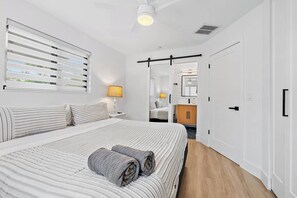 Une chambre à coucher avec un grand lit, une table de chevet, une lampe, une armoire et une salle de bain visible à travers une porte ouverte.