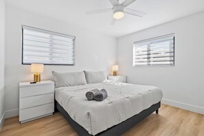 Une chambre moderne avec un lit, deux tables de chevet avec des lampes, un ventilateur de plafond et de grandes fenêtres équipées de stores.