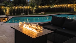 Un área junto a la piscina con chimenea, asientos exteriores y vista al agua.
