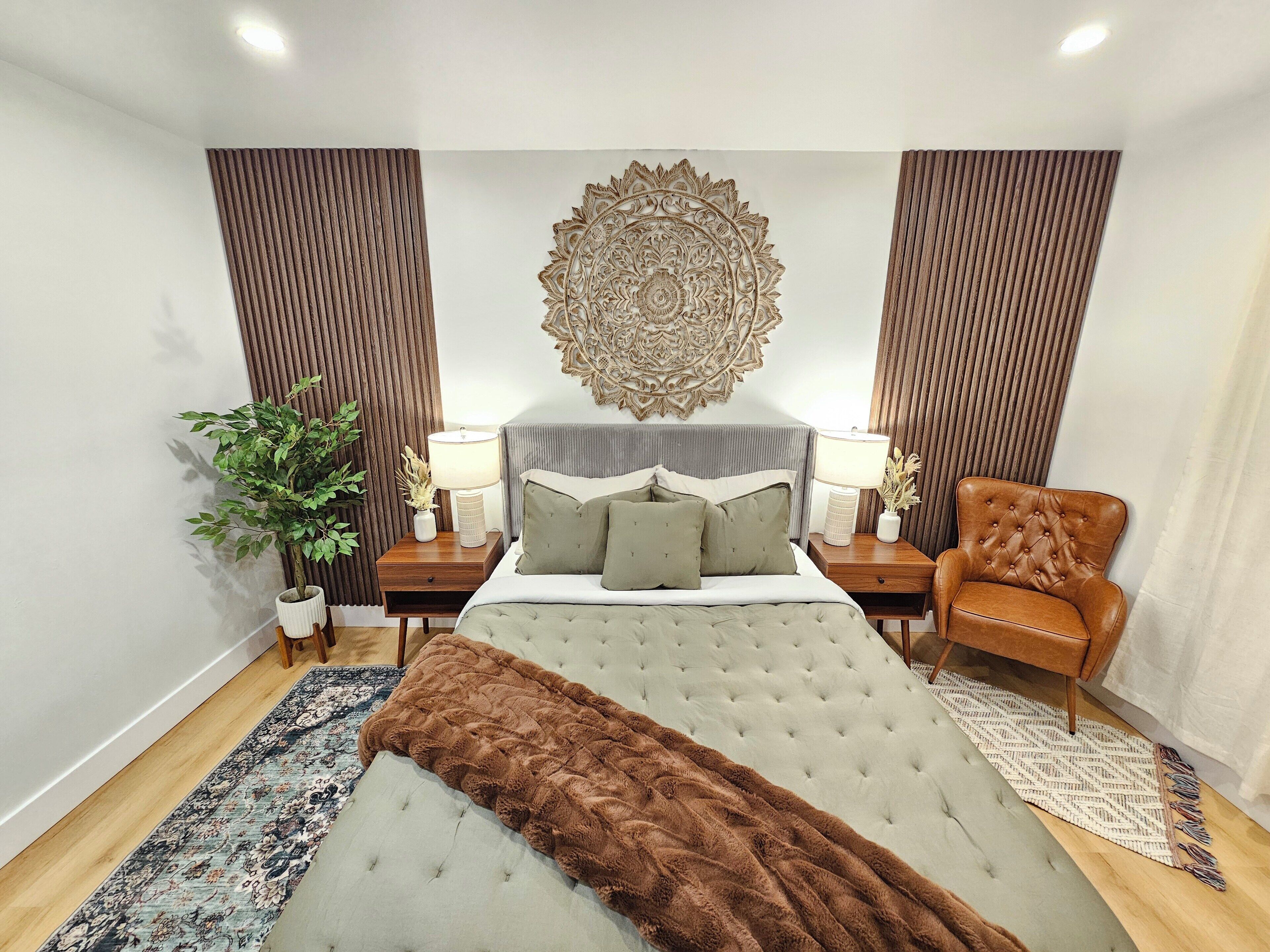 Une chambre à coucher avec un lit capitonné, deux tables de chevet avec des lampes, un élément décoratif mural, une plante en pot et un fauteuil marron.