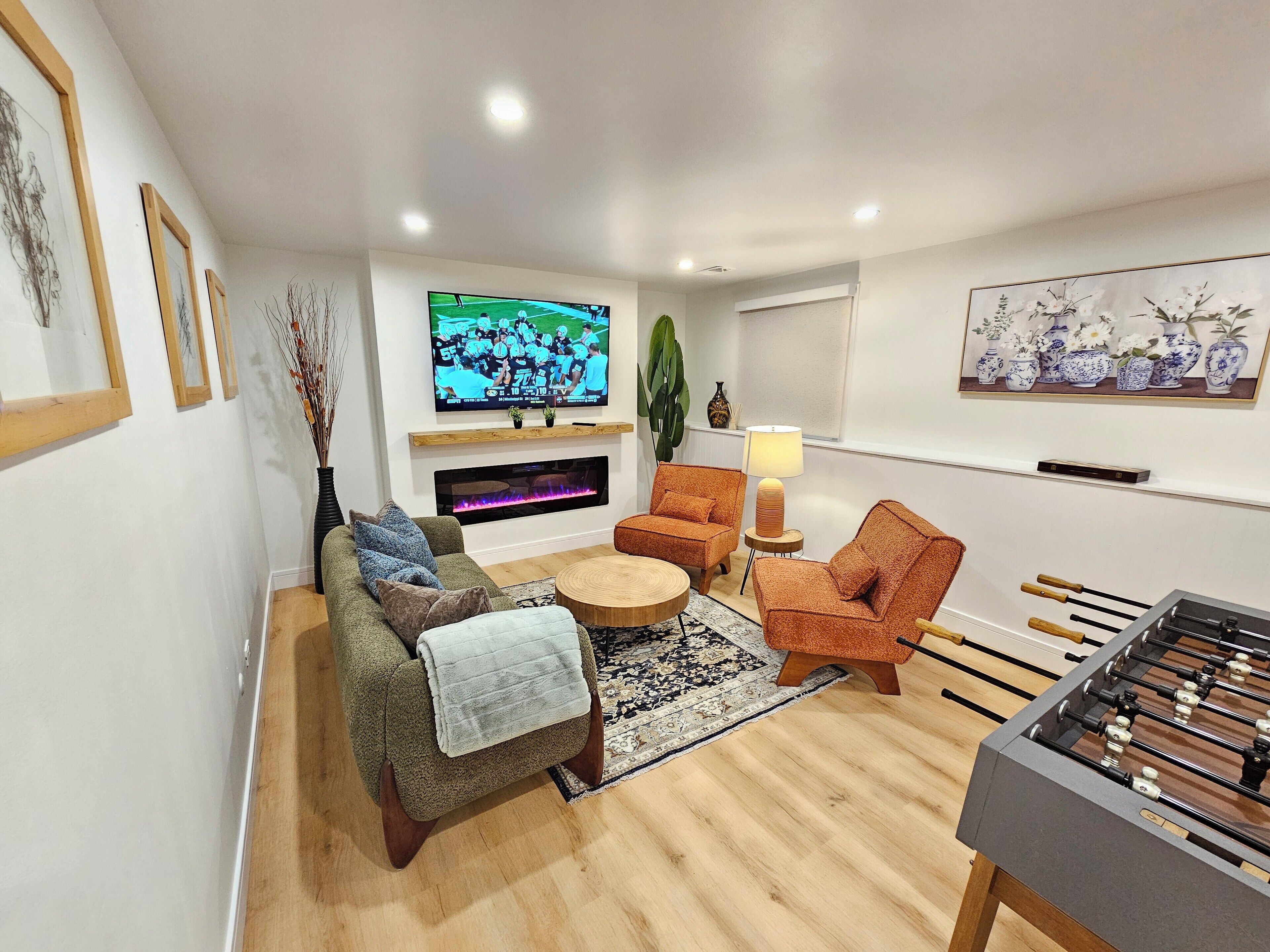 Un salon moderne comprenant un canapé, des fauteuils, une table basse, un téléviseur, une cheminée, une lampe et une table de baby-foot.