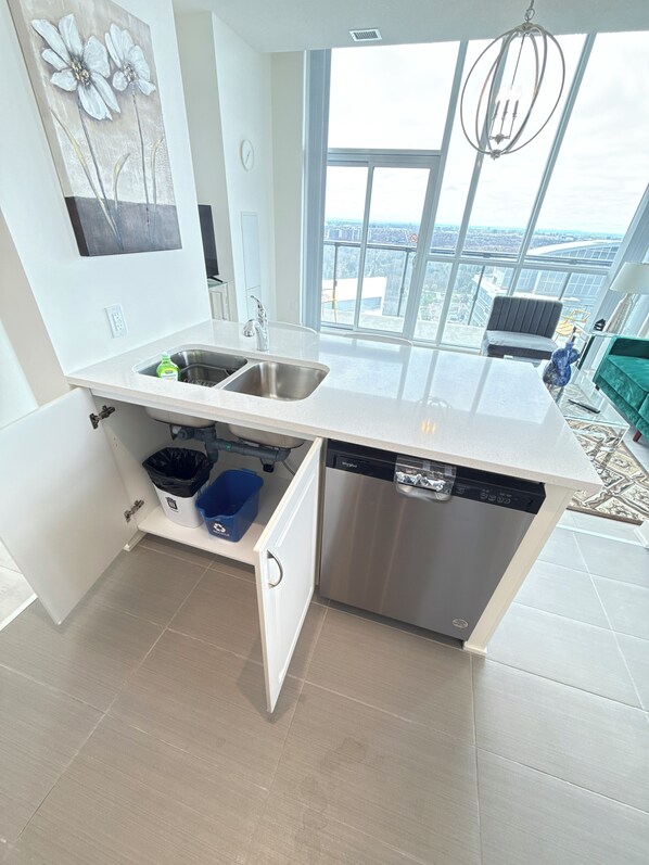 A modern kitchen with a dishwasher, double sink, and a view of the city through large windows.