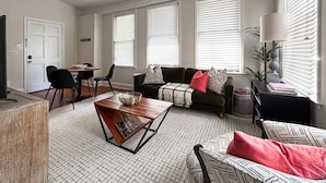 Ein Wohnzimmer mit Sofa, Couchtisch und Sesseln. Es gibt einen Esstisch mit Stühlen. Das Zimmer hat große Fenster mit Jalousien.