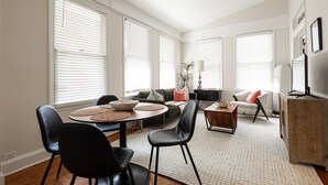 Ein geräumiges Wohnzimmer mit Essbereich, großen Fenstern und einem Fernseher.