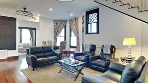 Ein geräumiges Wohnzimmer mit einer großen Sofagarnitur, einem Couchtisch und einem Massagesessel. Zwei Fenster mit Vorhängen, eine Tür und eine Lampe auf einem Beistelltisch sind ebenfalls vorhanden.