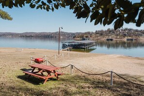 Ein Picknicktisch und ein Grill an einem sandigen Strand, im Hintergrund ein Steg und Boote.