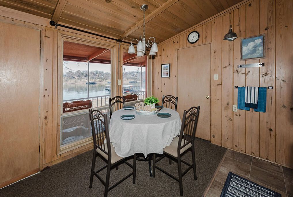 Una habitación con paneles de madera, una mesa redonda para cuatro personas, un reloj en la pared y vistas a un cuerpo de agua a través de una ventana.
