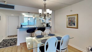 Un'area pranzo moderna con tavolo in vetro e sedie bianche, una cucina sullo sfondo e un quadro appeso al muro.