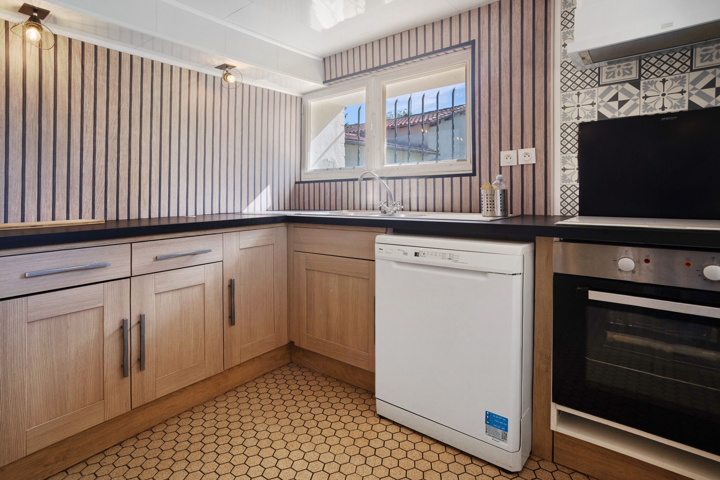 Uma cozinha com armários de madeira, lava-louças, forno, janela e chão revestido de azulejos.