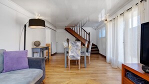 A modern living room with a sofa, a dining table with chairs, a staircase, and a television on a wooden stand.