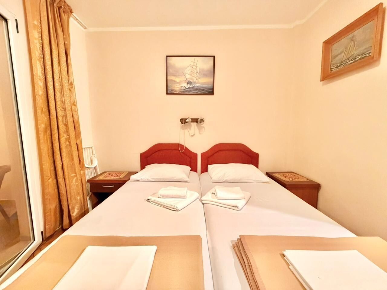 A hotel room with two beds, a window with curtains, a painting on the wall, and a nightstand with a lamp.