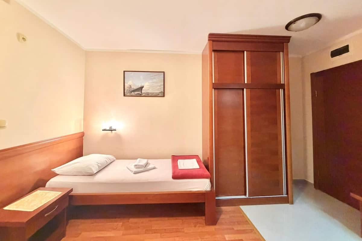 A small, single-room hotel room with a wooden bed, a nightstand, a wardrobe, and a wall-mounted light.